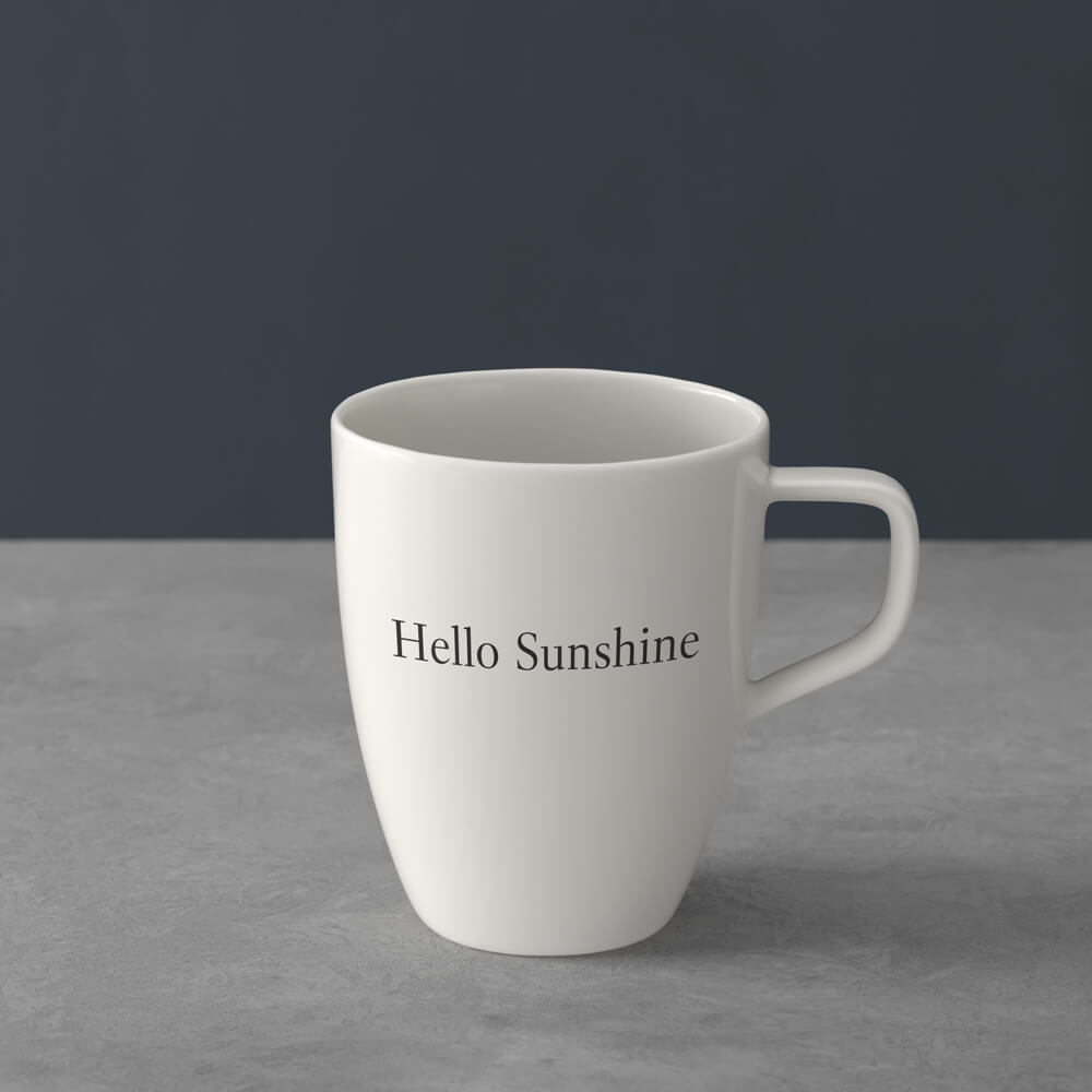 1041309651_villeroy&boch_schwarz Weiße Keramiktasse mit dem Aufdruck "Hello Sunshine" auf einer grauen Fläche vor einem dunklen Hintergrund.