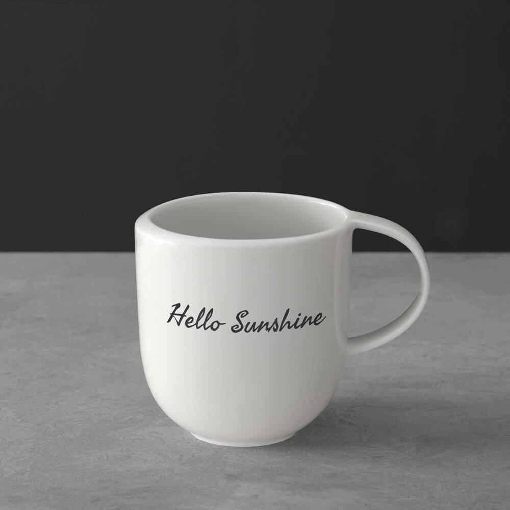 1042649651_Seaweed-Skript_schwarz Weiße Keramiktasse mit dem Schriftzug "Hello-Sunshine" in schwarzer Schrift auf einer grauen Fläche vor dunklem Hintergrund.