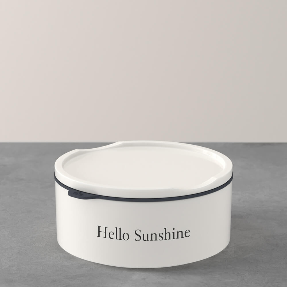 1048699410_Villeroy-Boch_schwarz Auf einer grauen Fläche steht ein weißer, runder Behälter mit Deckel, auf dessen Vorderseite die Aufschrift "Hello Sunshine" steht.