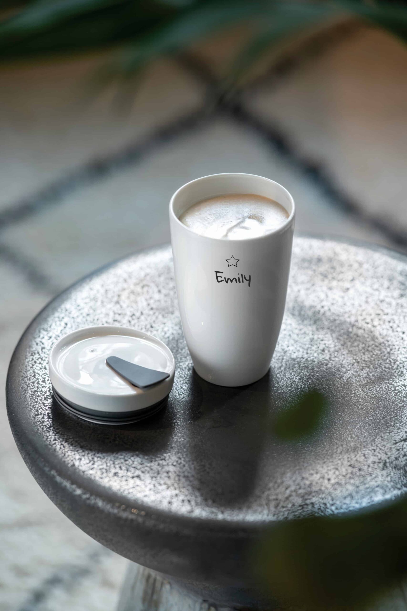 Ein weißer Reisebecher mit der Aufschrift "Emily" und einem Deckel daneben steht auf einem runden, gesprenkelten Tisch.
