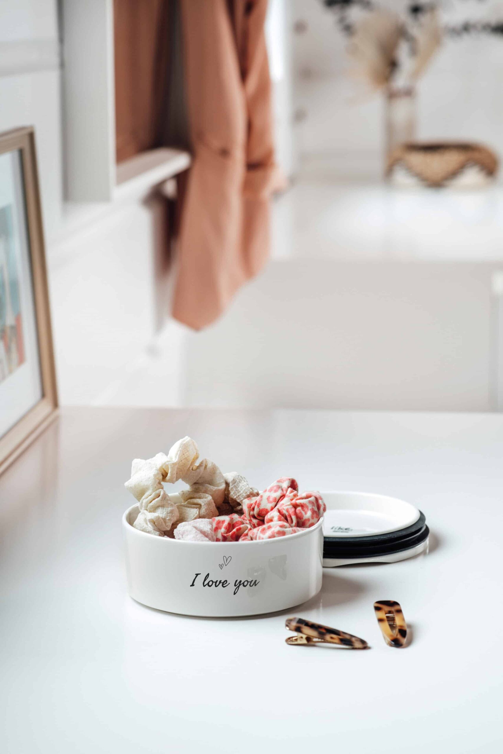 1048699410_ToGo&ToStay_mothersday_1 Eine weiße Schale mit der Aufschrift "Ich liebe dich" enthält Haargummis und steht auf einem weißen Schreibtisch, neben dem Haarspangen liegen.