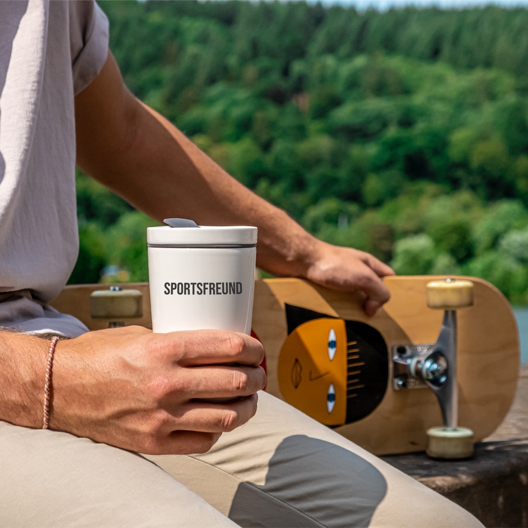 Eine Person mit einem SPORTSFEUND-Reisebecher sitzt im Freien und hat ein Skateboard auf dem Schoß