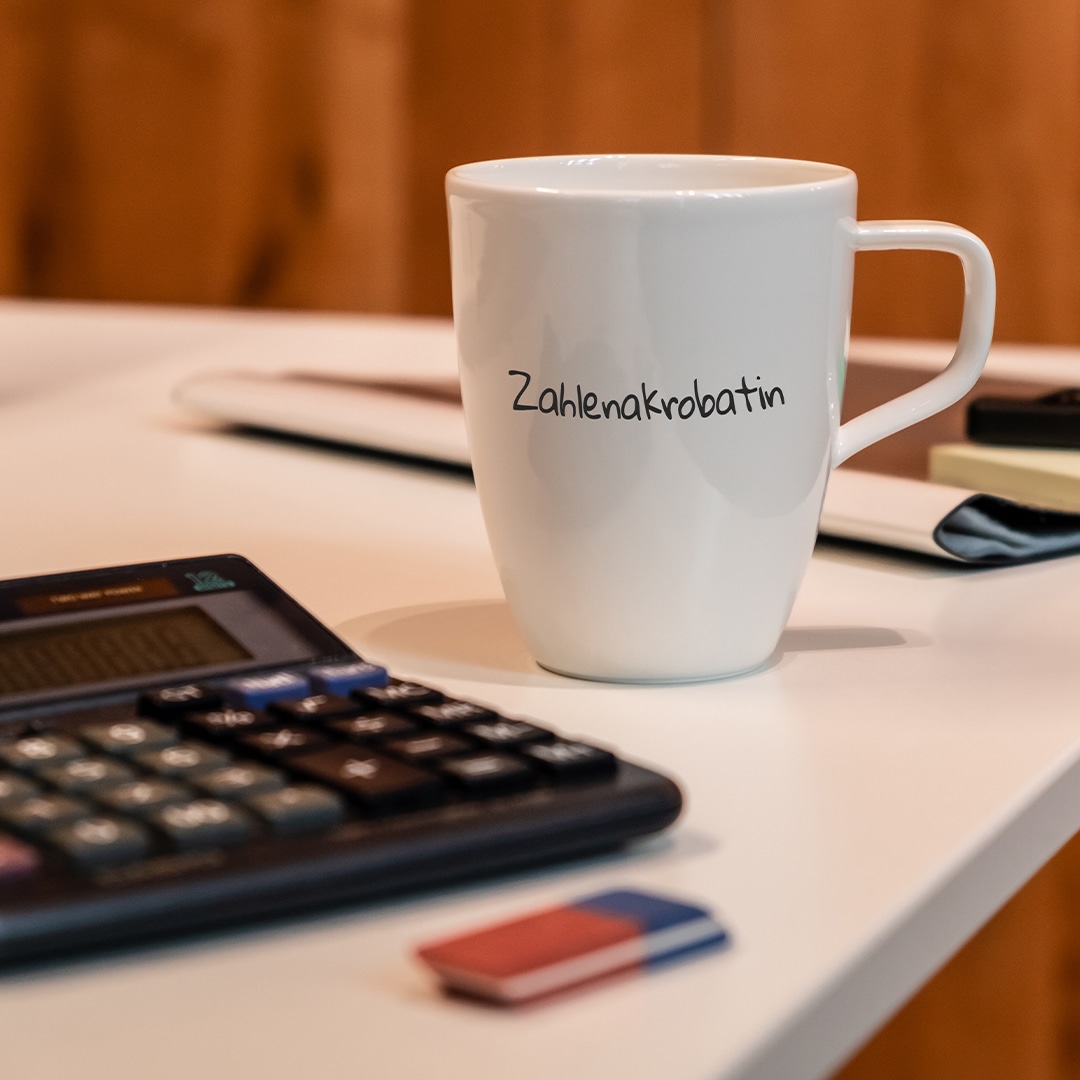 Image_A_2 Ein weißer Becher mit der Aufschrift "Zahlenakrobatin" steht auf einem Schreibtisch neben einem Taschenrechner, einem Stift und einem Radiergummi.