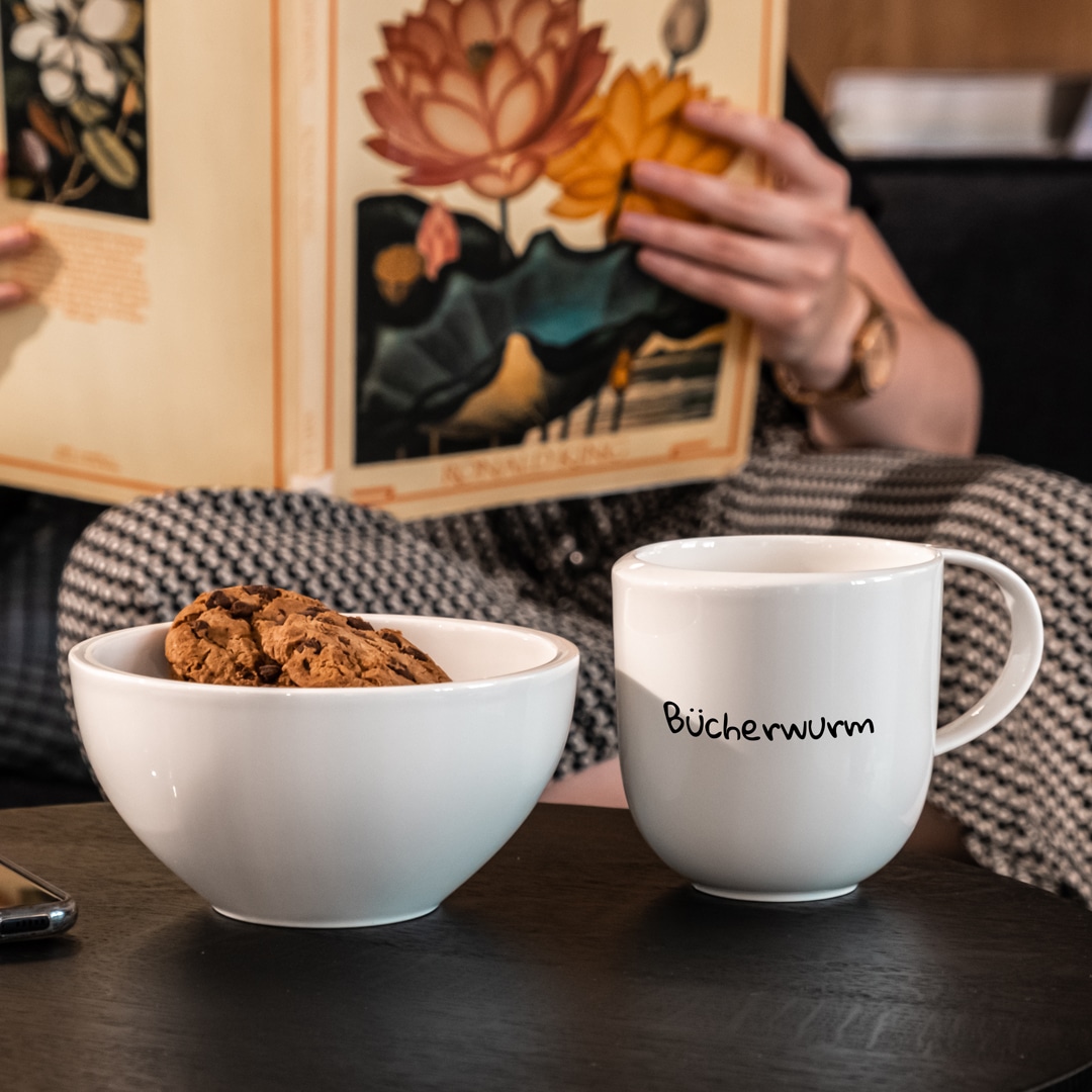Image_B Eine Person, die ein Buch liest, sitzt hinter einer Schüssel mit Keksen und einem weißen Becher mit der Aufschrift "Bücherwurm" auf einem Tisch.