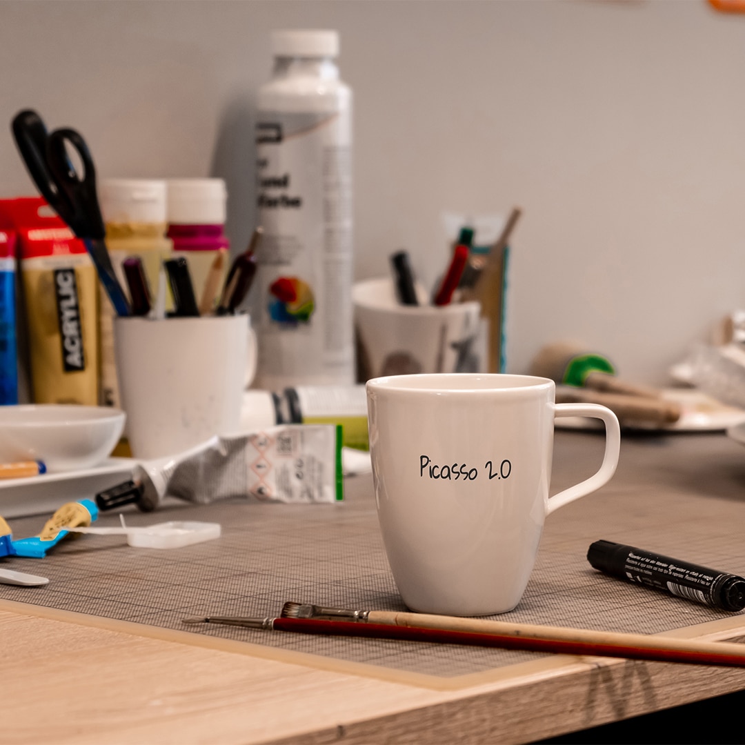 Image_B_1 Ein weißer Becher mit der Aufschrift "Picasso 2.0" steht auf einem vollgestopften Kunstschreibtisch mit Pinseln, Farben und Kunstzubehör.