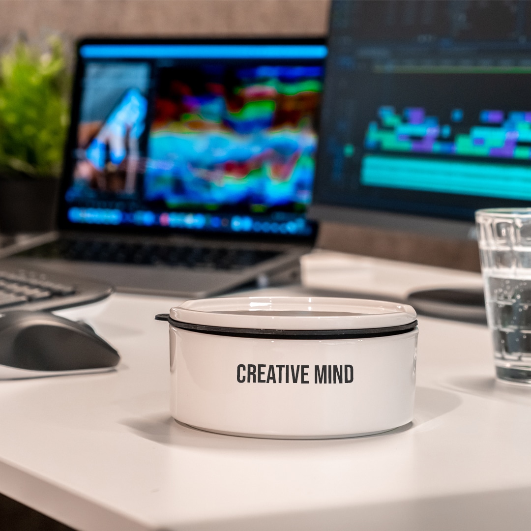 Image_C Weißer Behälter mit der Aufschrift "Creative Mind" auf dem Schreibtisch, daneben zwei Computermonitore, ein Glas Wasser und eine Maus.