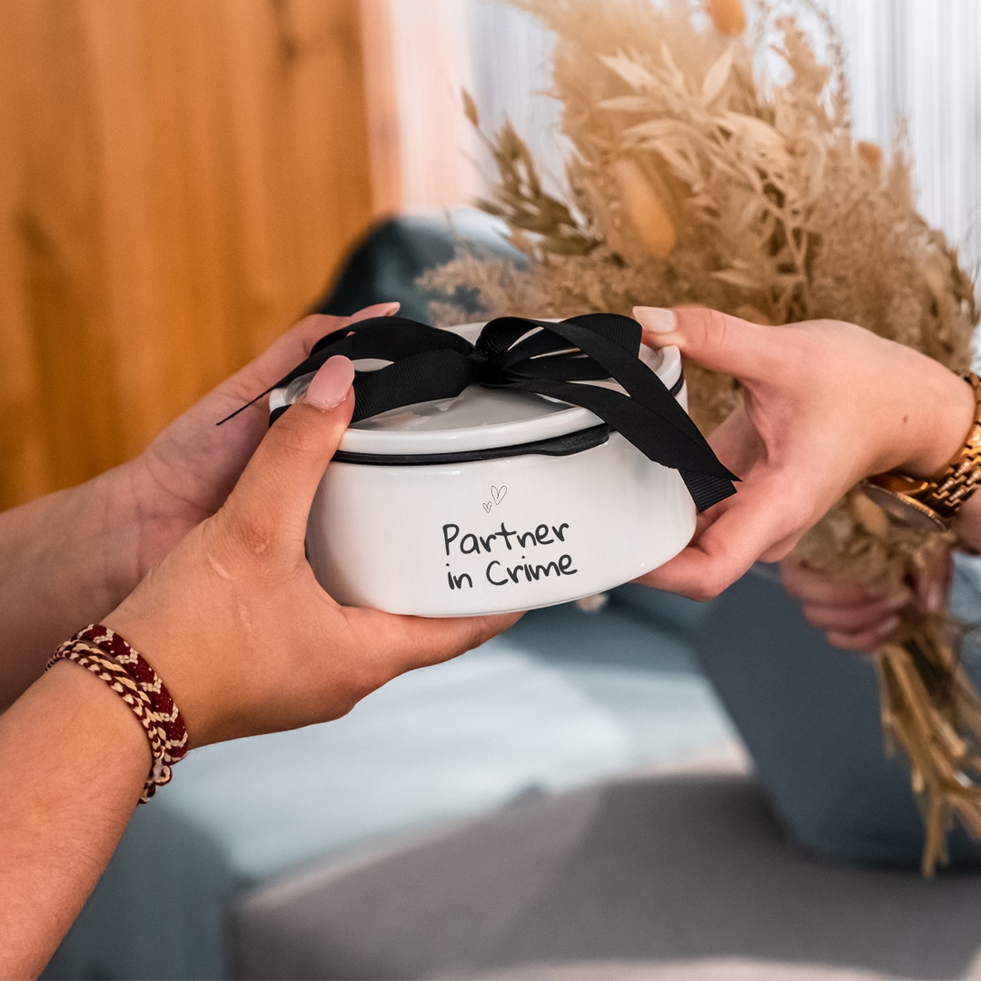 Image_C_1 Zwei Personen tauschen eine runde Geschenkbox mit der Aufschrift "Partner in Crime" aus, wobei eine Person einen Strauß getrockneter Blumen in der Hand hält.