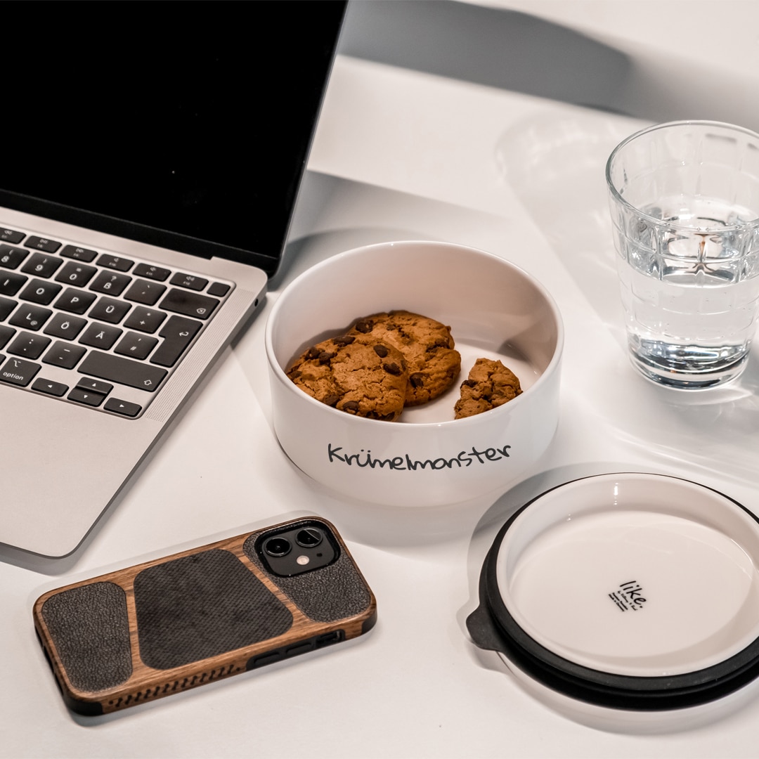 Image_D Eine Schale mit der Aufschrift "Krümelmonster" mit Keksen, ein Smartphone, ein Laptop, eine leere Schüssel und ein Glas Wasser auf einem weißen Tisch.