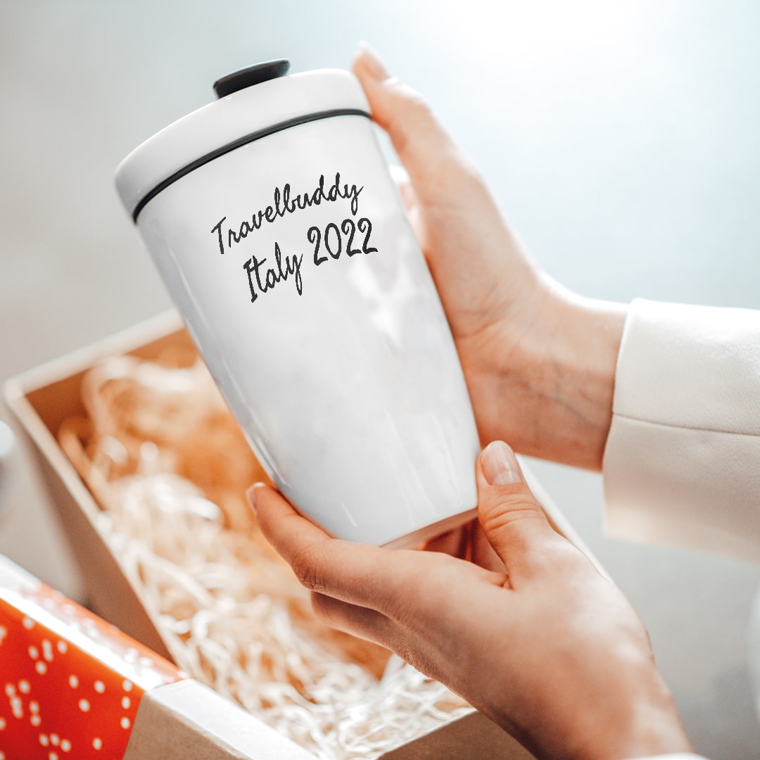 Eine Person hält einen weißen Reisebecher mit der Aufschrift "Travelladdy Italy 2022" neben einer offenen, mit Stroh gefüllten Geschenkbox.