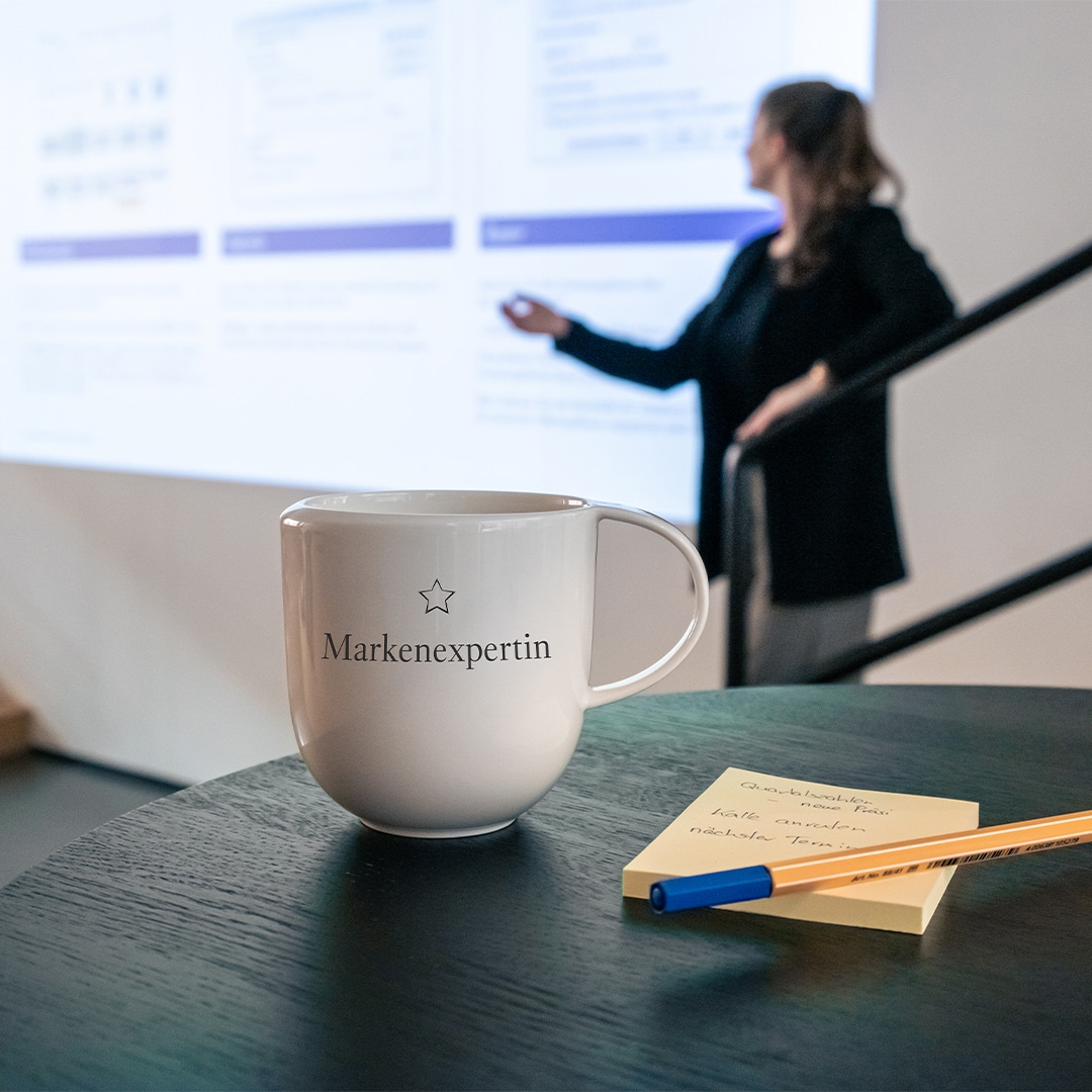 Image_E Weißer Becher mit der Aufschrift "Markenexpertin" auf einem Tisch mit einem Stift, einem Notizblock und einer Frau, die im Hintergrund etwas präsentiert.