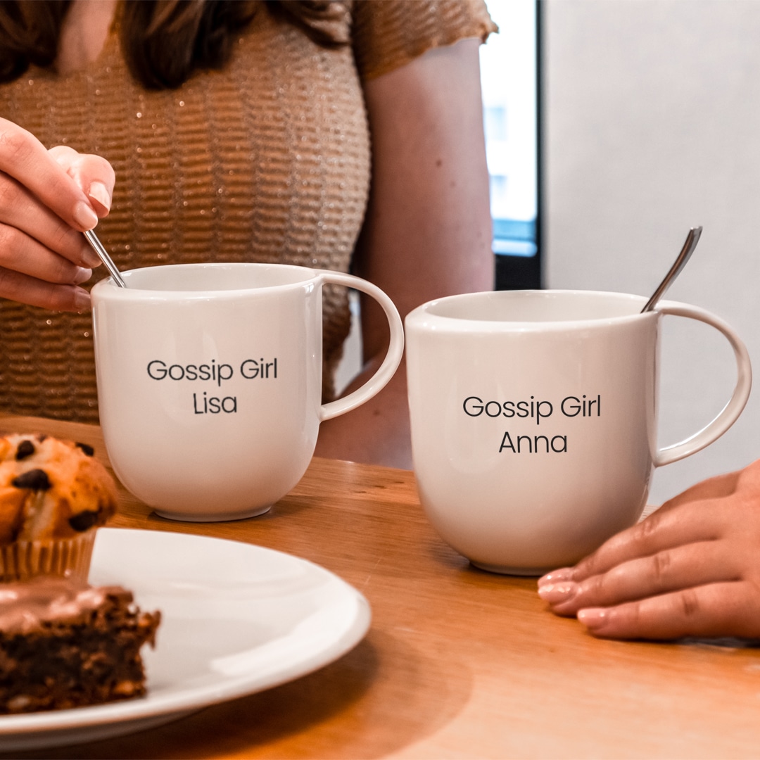 Image_E_1 Zwei Frauen sitzen an einem Tisch mit Tassen mit der Aufschrift "Gossip Girl Lisa" und "Gossip Girl Anna", daneben stehen Muffins und Kuchen.