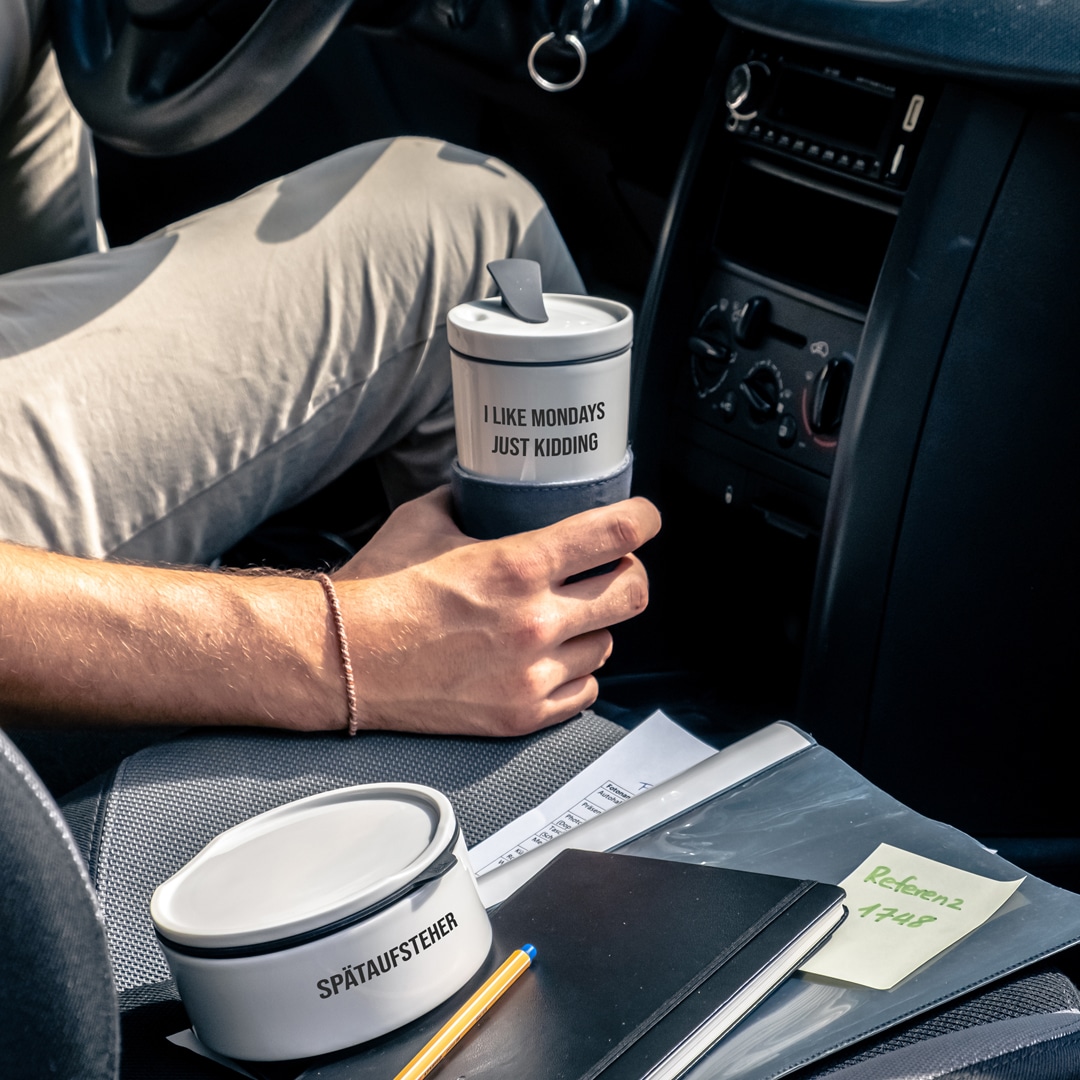 Eine Person in einem Auto hält einen Becher mit der Aufschrift "I LIKE MONDAYS JUST KIDDING", auf dem Sitz liegen Notizbücher und ein Stift.