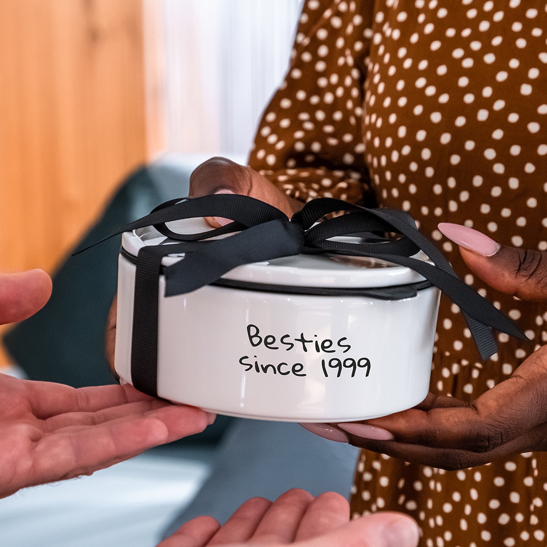 Image_F_2 Eine Person überreicht einer anderen Person eine weiße Geschenkschachtel mit der Aufschrift "Besties since 1999" und einer schwarzen Schleife.