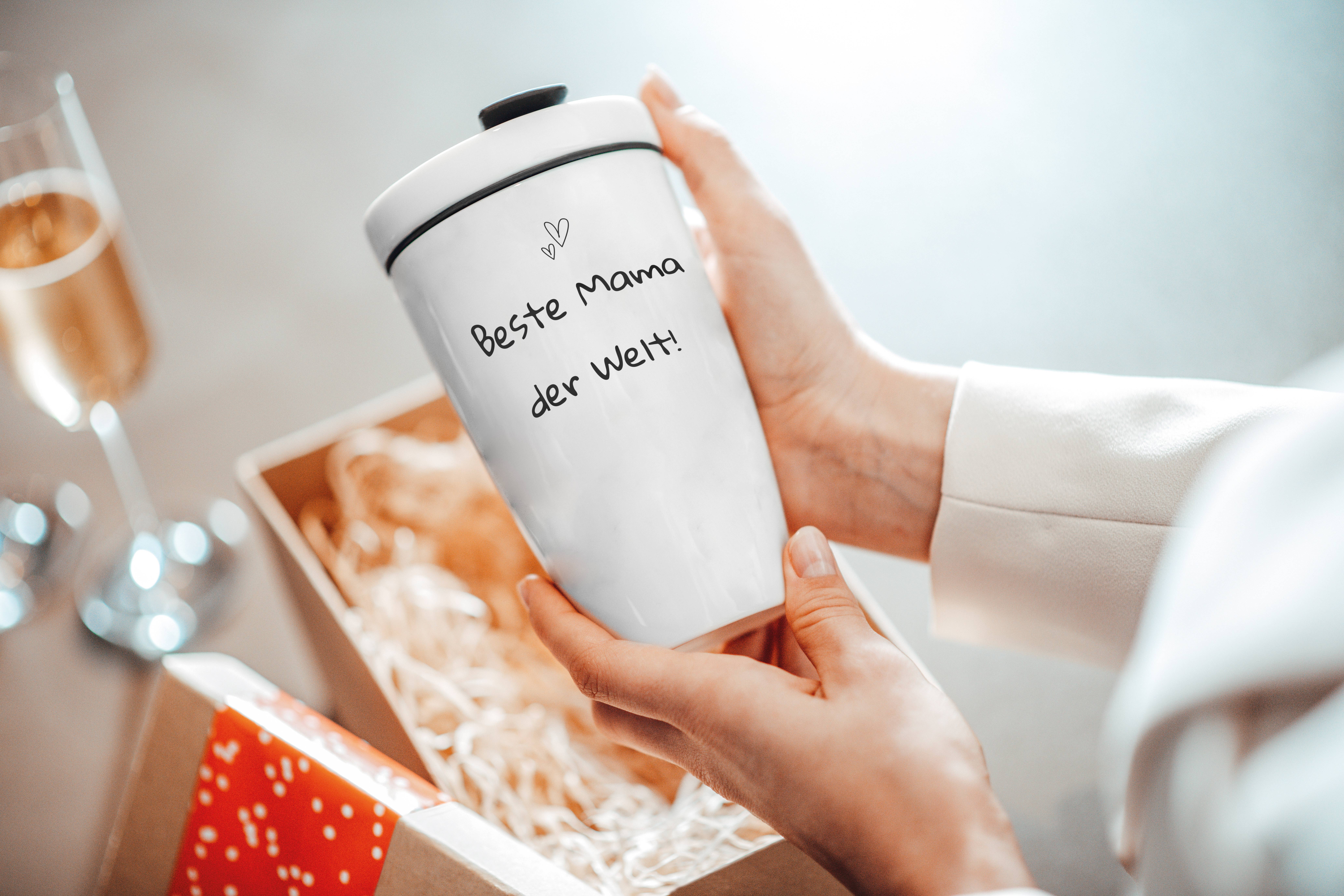 Eine Person hält einen weißen Becher mit der Aufschrift "Beste Mama der Welt" in der Hand, daneben eine Geschenkbox und ein Glas Sekt.
